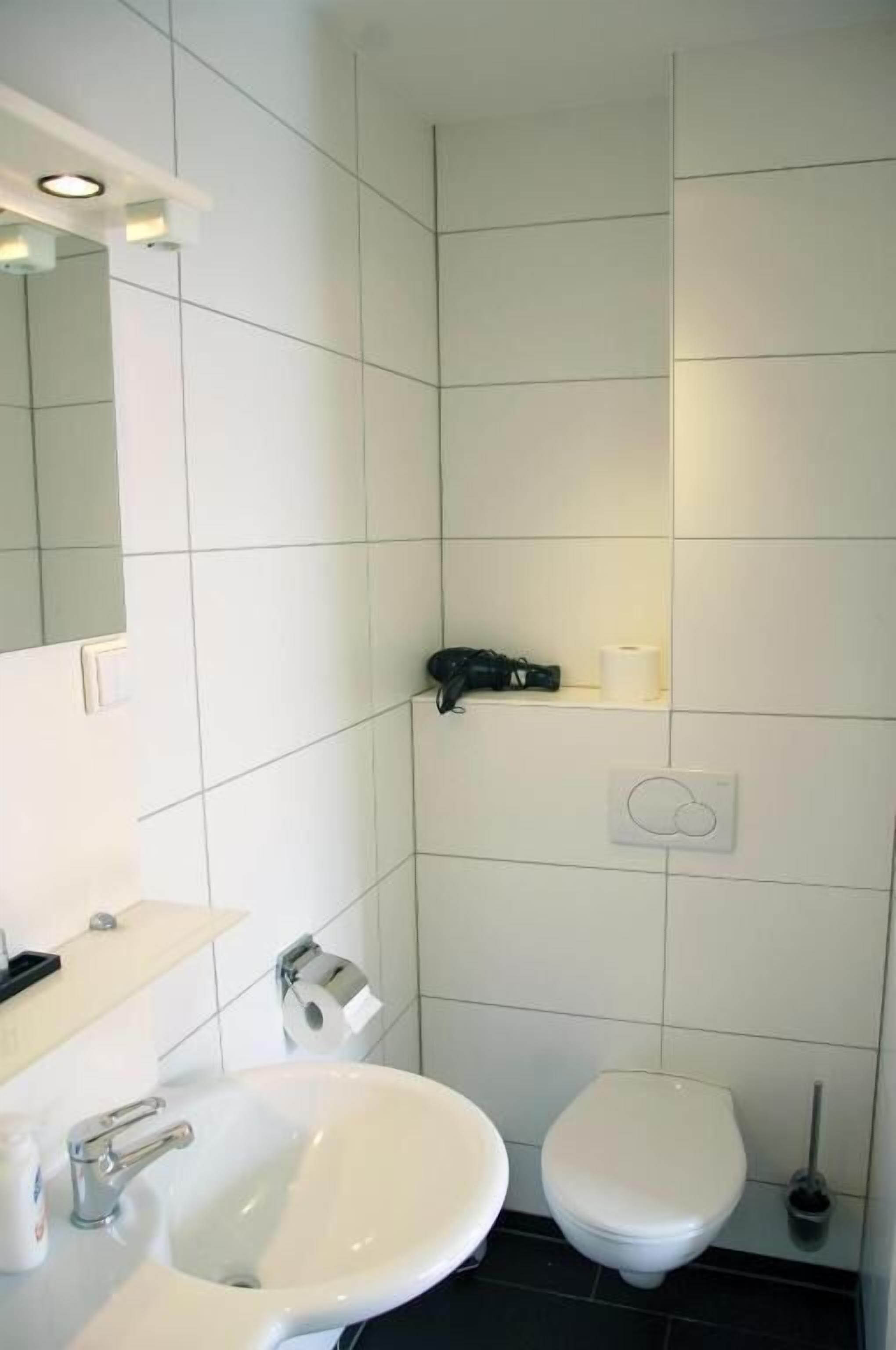 Un baño con baldosas blancas, un lavamanos, un inodoro, un espejo y un estante con artículos de aseo.