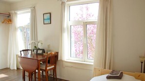 Ein Zimmer mit Esstisch und Stühlen aus Holz, einem Bett mit weißer Bettdecke und einem Fenster mit bunten Blumen davor.