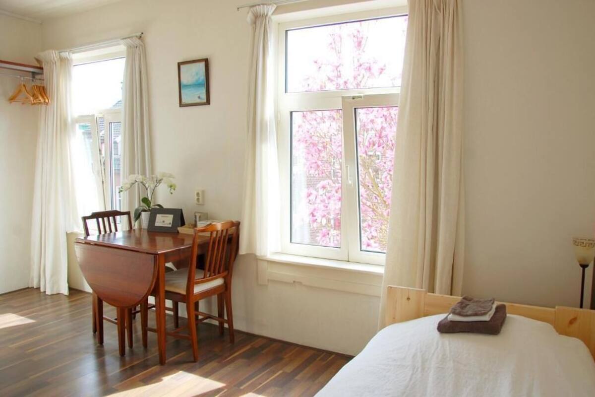 A room with a wooden dining table and chairs, a bed with a white comforter, and a window with pink flowers outside.