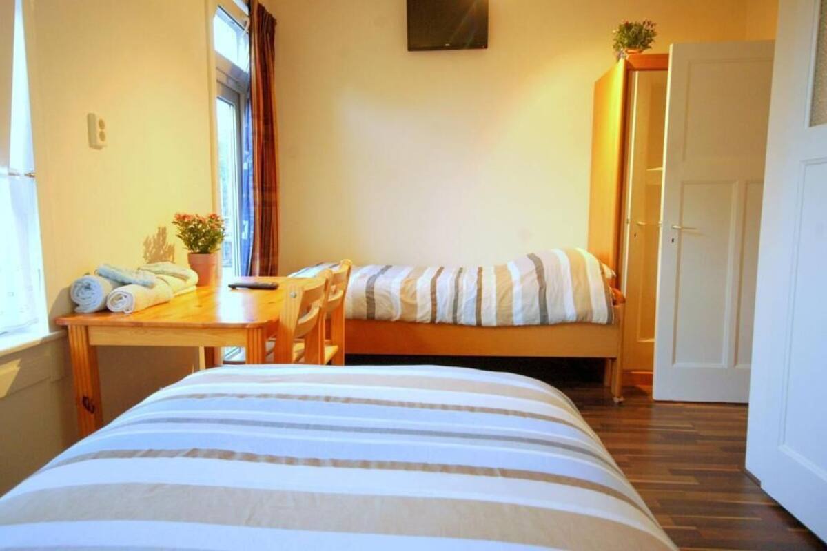 A small, cozy room with a bed, a wooden table, a chair, and a window with curtains.