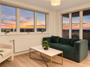 A modern living room with a green sofa, a marble coffee table, and large windows with a sunset view.