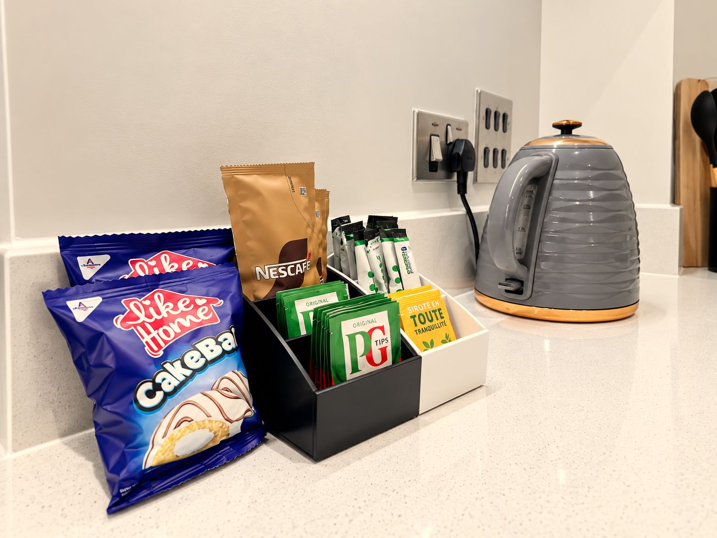 A kitchen counter with a coffee maker, a bag of Nescafe coffee, a box of tea bags, a bag of cake mix, and a box of coffee tips.