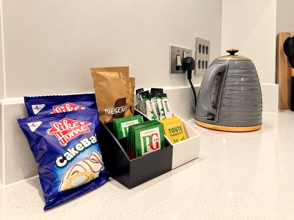 A kitchen counter with a coffee maker, a bag of Nescafe coffee, a box of tea bags, a bag of cake mix, and a box of coffee tips.
