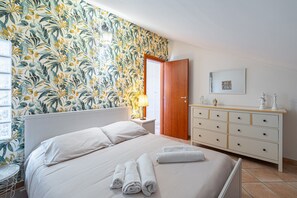 Une chambre à coucher comprenant un lit, une table de chevet, une commode, une porte et un mur recouvert d’un papier peint à motifs de feuilles.