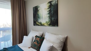 Ein Hotelzimmer mit Bett, weißen Kissen, einem Wandbild mit Waldmotiv und Blick ins Freie durch ein Fenster mit Vorhängen.