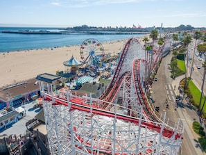 Una montaña rusa en un parque de atracciones frente al mar, con una playa y muelle al fondo.