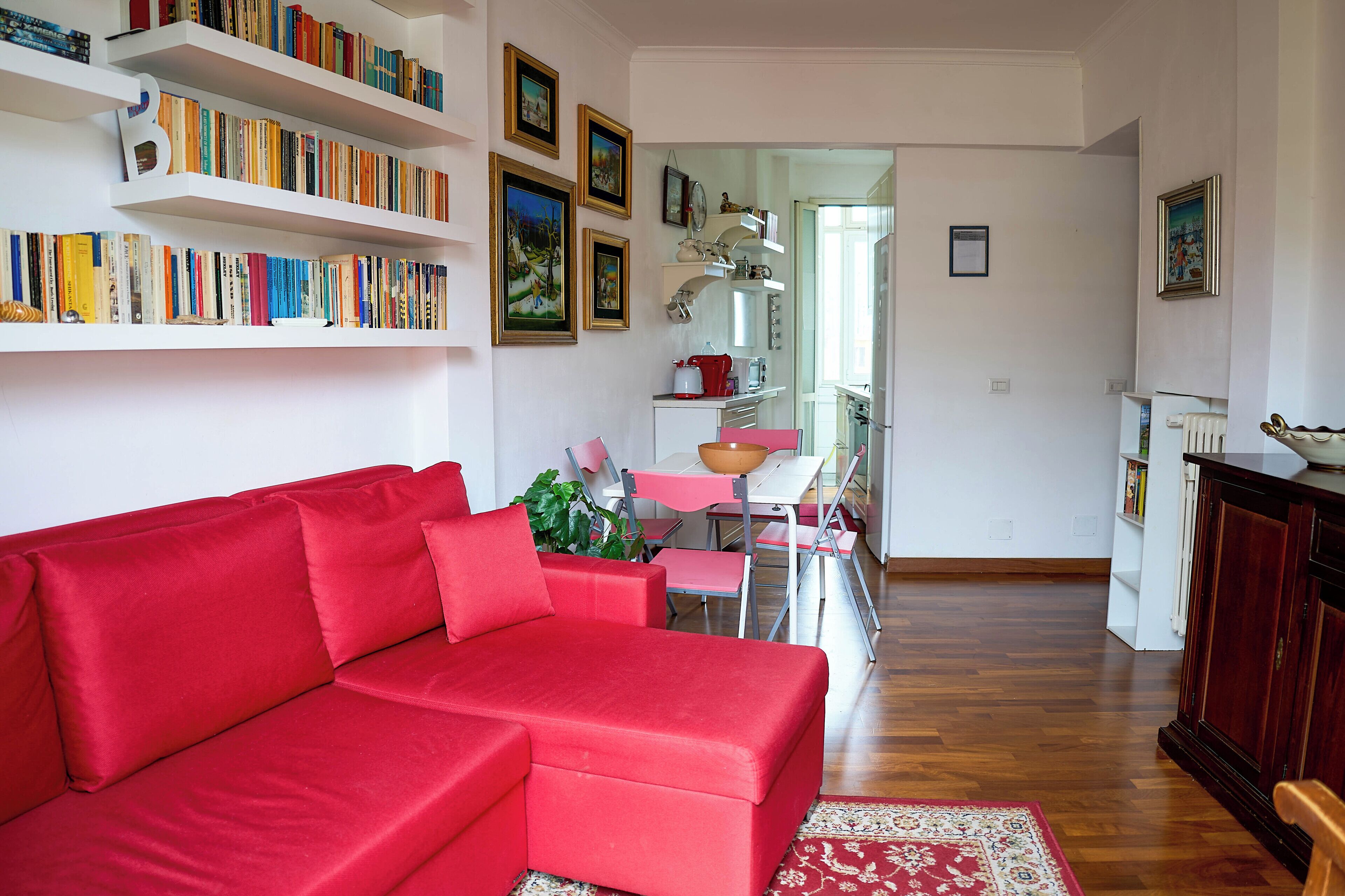 A living room with a red sofa, a bookshelf filled with books, and a dining area with pink chairs and a table.