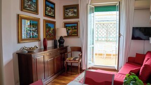 A living room with a red sofa, wooden cabinet, and a chair. There are framed pictures on the wall and a window with curtains. - Naif (Rome)