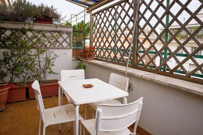 Uma área de pátio com mesa e cadeiras brancas, plantas em vasos e vista para prédios através de uma janela com treliça.