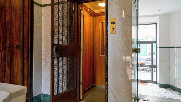A hotel lobby with a wooden door, a glass elevator, and a marble floor. - Naif (Rome)