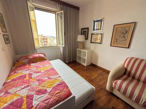 Quarto de hotel com cama, uma cadeira, uma mesinha e janela com vista para edifícios.