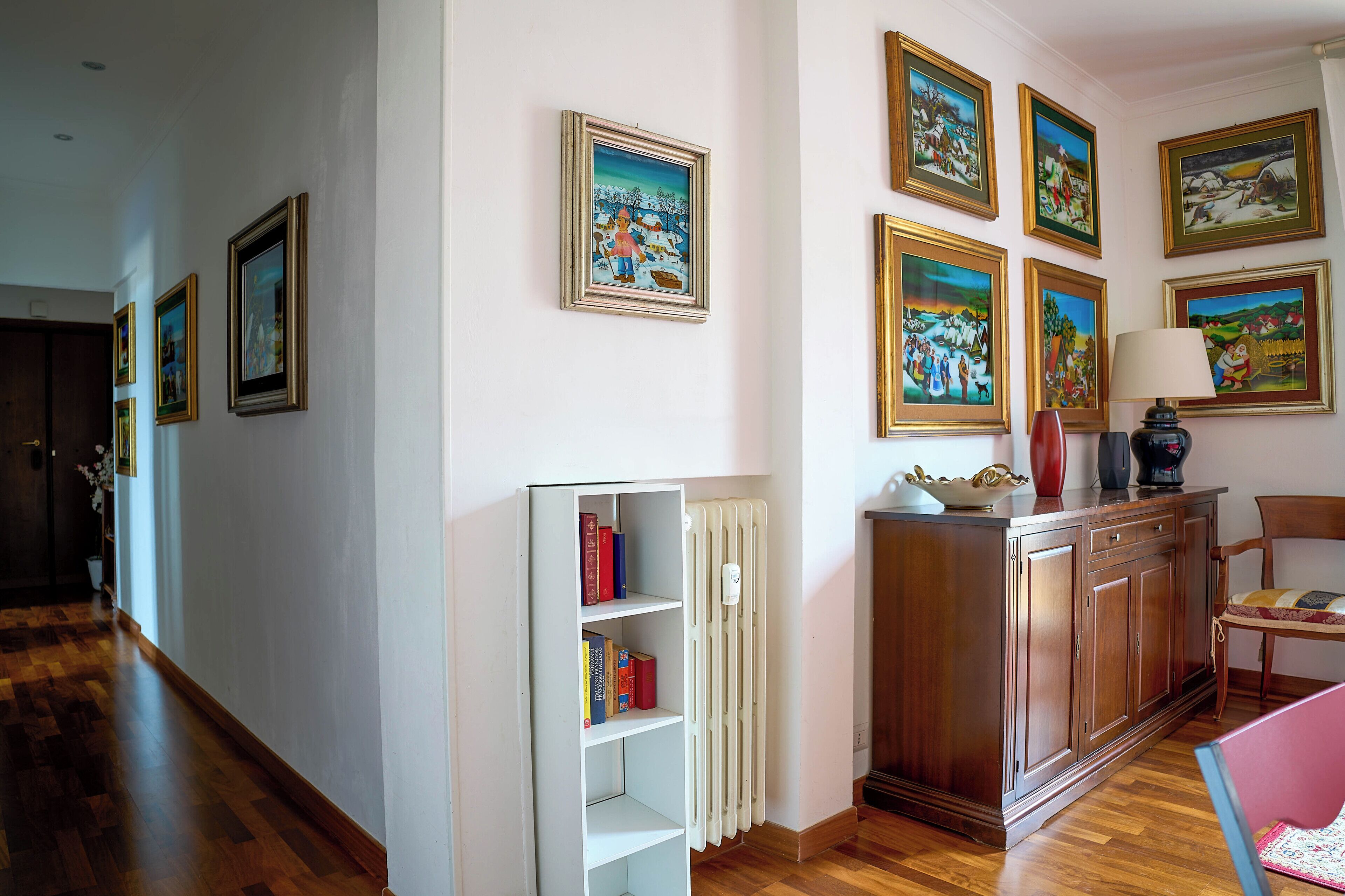A hallway with wooden flooring, a radiator, a bookshelf, and a cabinet with a lamp.