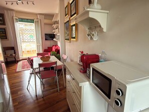 Uma cozinha compacta com micro-ondas, máquina de café e torradeira vermelha. Há uma mesa de jantar com cadeiras e um sofá vermelho na área de estar adjacente.
