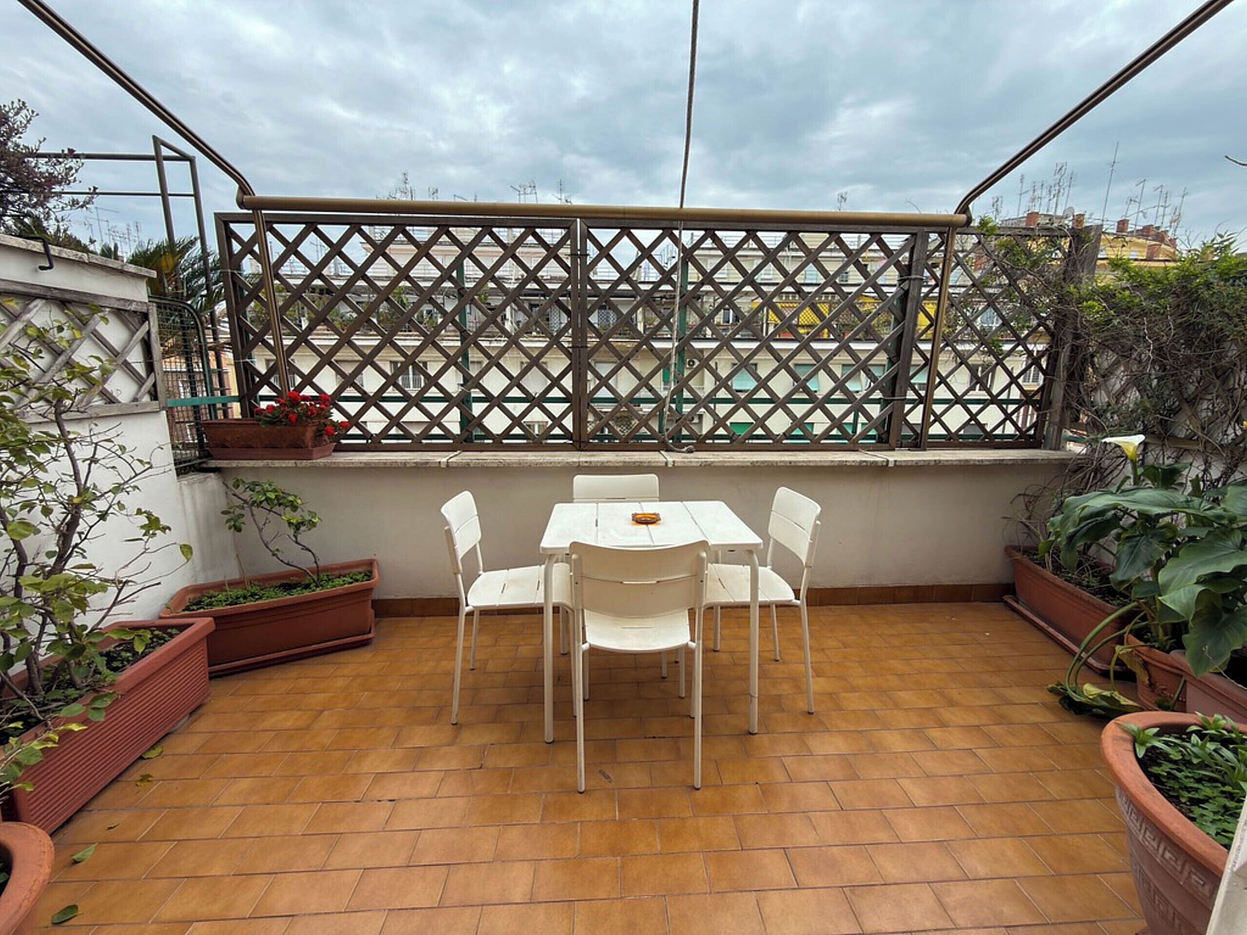 Eine Terrasse mit Tisch und Stühlen, umgeben von Kübelpflanzen und einem Gitterzaun.