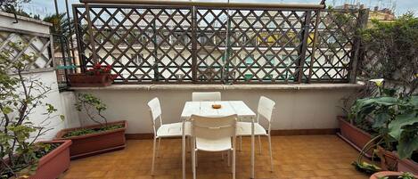 Eine Terrasse mit Tisch und Stühlen, umgeben von Kübelpflanzen und einem Gitterzaun.