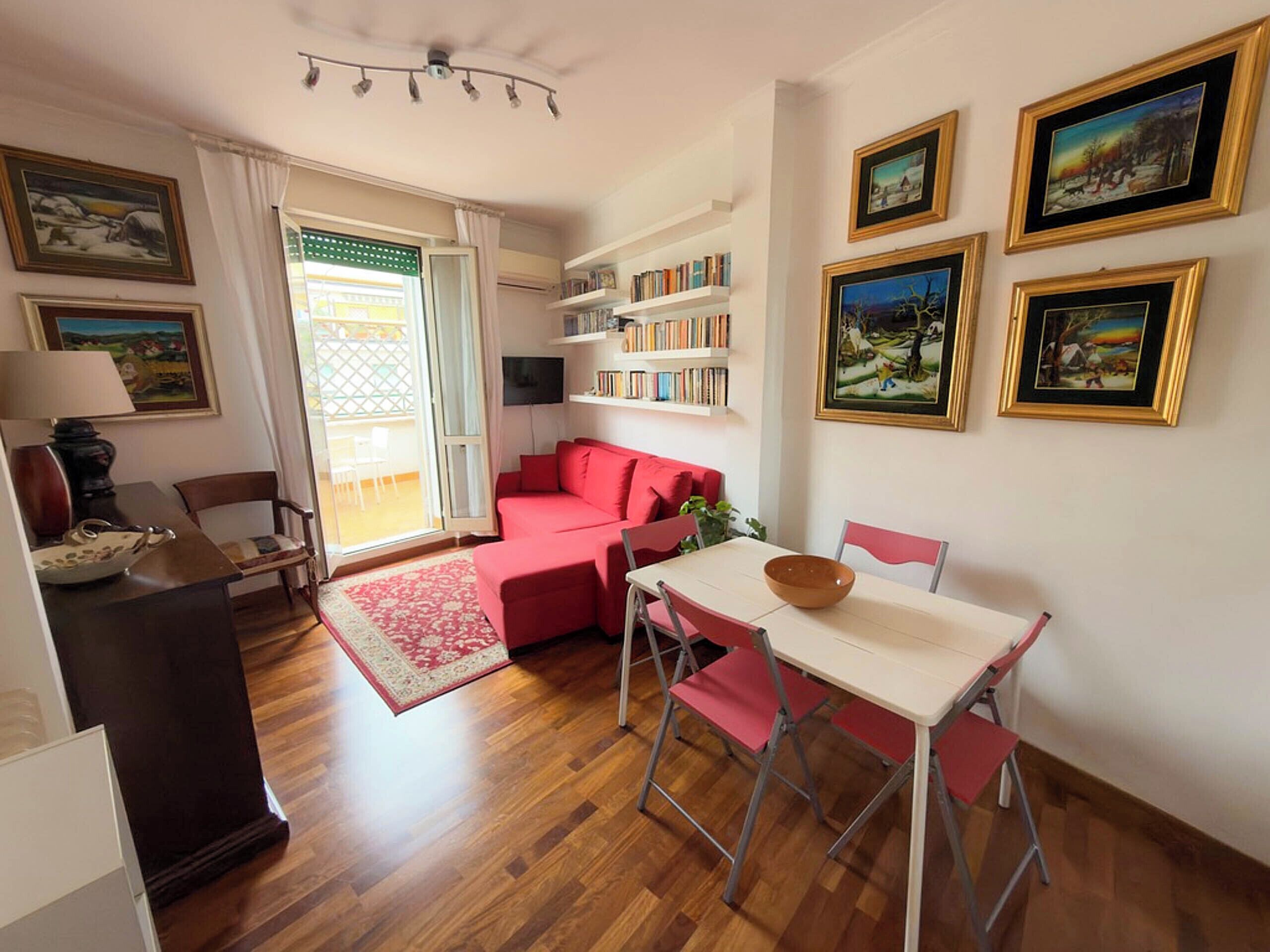 A cozy living room with a red sofa, a dining table with chairs, a bookshelf, and a TV mounted on the wall.
