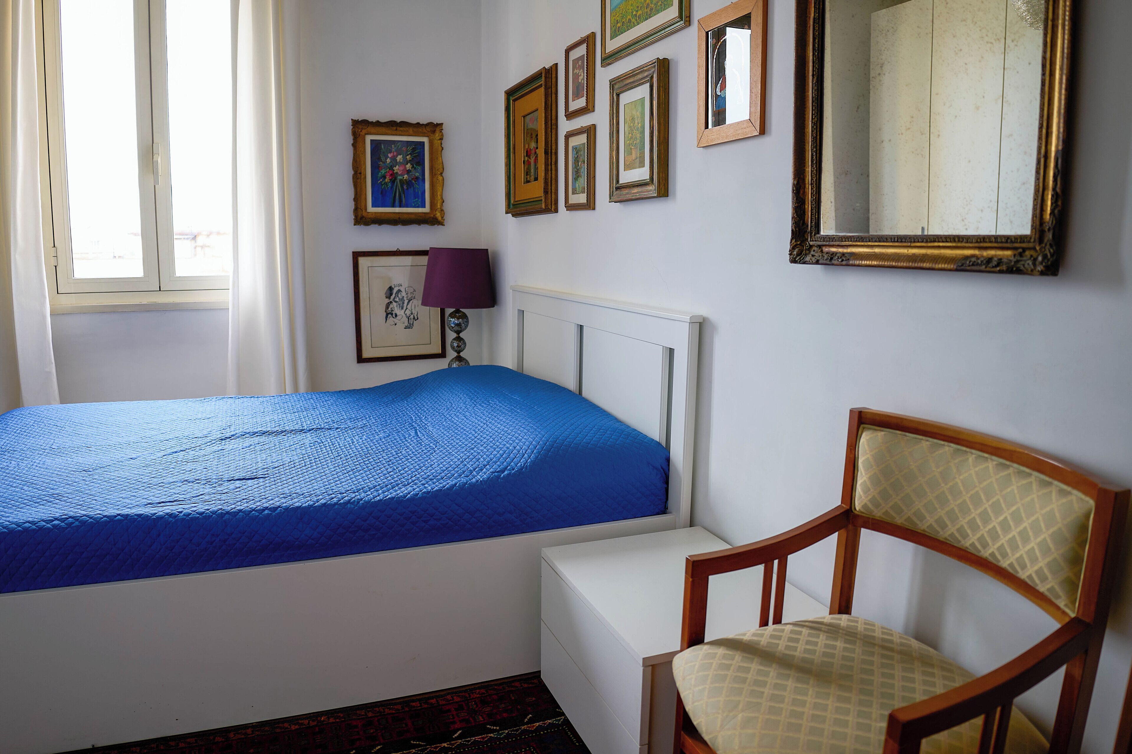 A bedroom with a bed, a chair, a bedside table, a lamp, and several framed pictures on the wall.