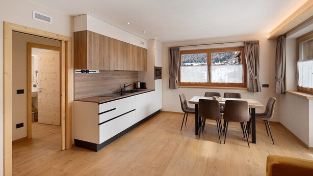 A modern kitchen with wooden cabinets, a dining area with a table and chairs, and a view of a snowy landscape through a window. - Chalet 'Arnica' with Mountain View, Balcony and Wi-Fi (Livigno)