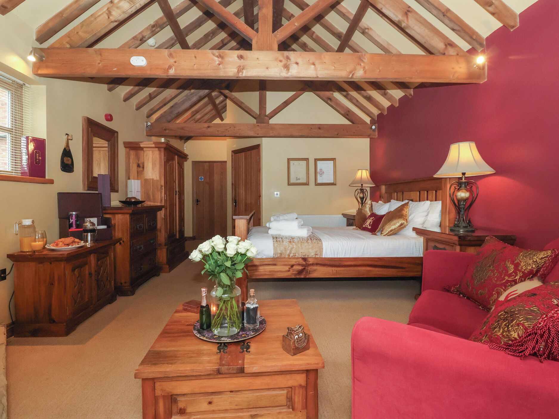Una habitación con una cama, una mesa de madera con un florero y un sofá rojo, además de una estructura de techo de madera.