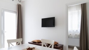Ein modernes Wohnzimmer mit einem Flachbildfernseher an der Wand, einem Esstisch mit Stühlen und einem kleinen Beistelltisch mit einem dekorativen Gegenstand.