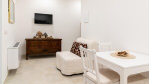 Ein kleines, sauberes Zimmer mit einem weißen Tisch, einem weißen Sofa, einem Holzschrank und einem an der Wand befestigten Flachbildfernseher.