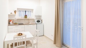 Eine kleine, helle Küche mit weißen Schränken, einer Mikrowelle und einem Kühlschrank. Es gibt einen Esstisch mit Stühlen und ein Fenster mit Vorhang.