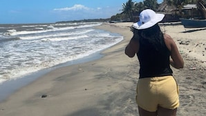 Una playa con cielo despejado, una persona con pantones amarillos y sombrero blanco, y un bote en la arena.