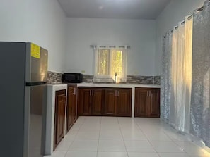 A kitchen with a refrigerator, microwave, wooden cabinets, a window with curtains, and a tiled floor.