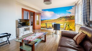 Een woonkamer met een televisie, een glazen salontafel, een bruine leren bank en uitzicht op een wijngaard door grote ramen.
