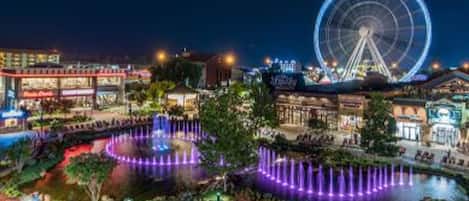 A nighttime view of a waterfront area with a large Ferris wheel, a fountain, and various buildings.