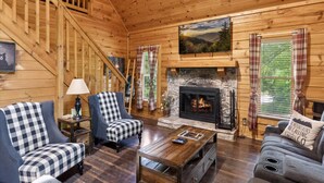 L’intérieur d’un chalet en bois, avec une cheminée, des canapés à carreaux, un escalier et un tableau accroché au mur.