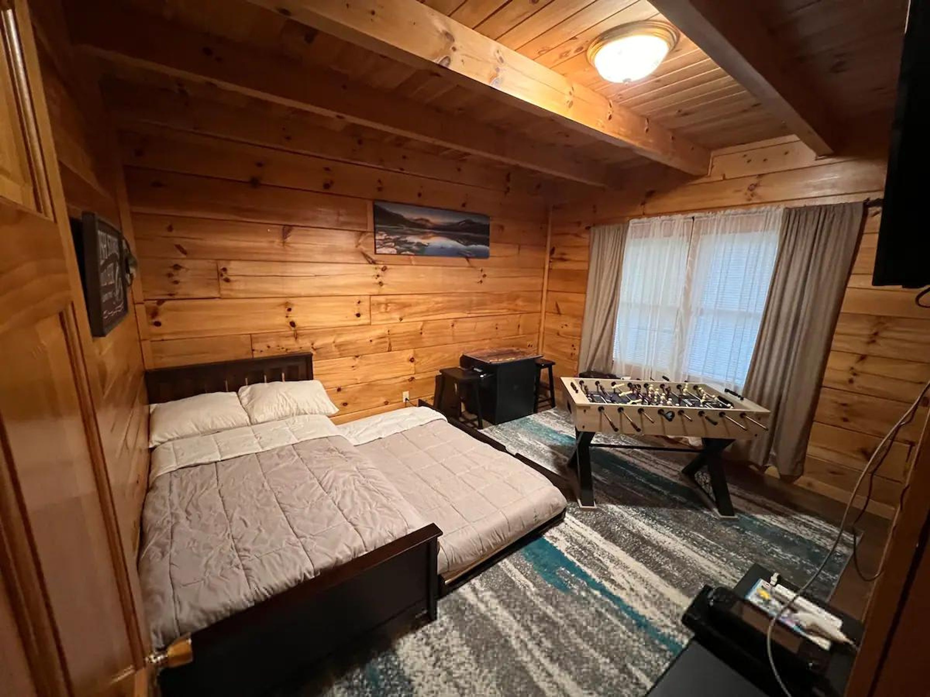 A wooden-paneled room with a bed, a foosball table, and a window with curtains.