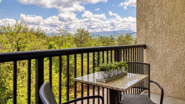 A balcony with a table and chairs, a railing, and a view of greenery and mountains. - Mountain View Condo by Beyond Expectations (Pigeon Forge)