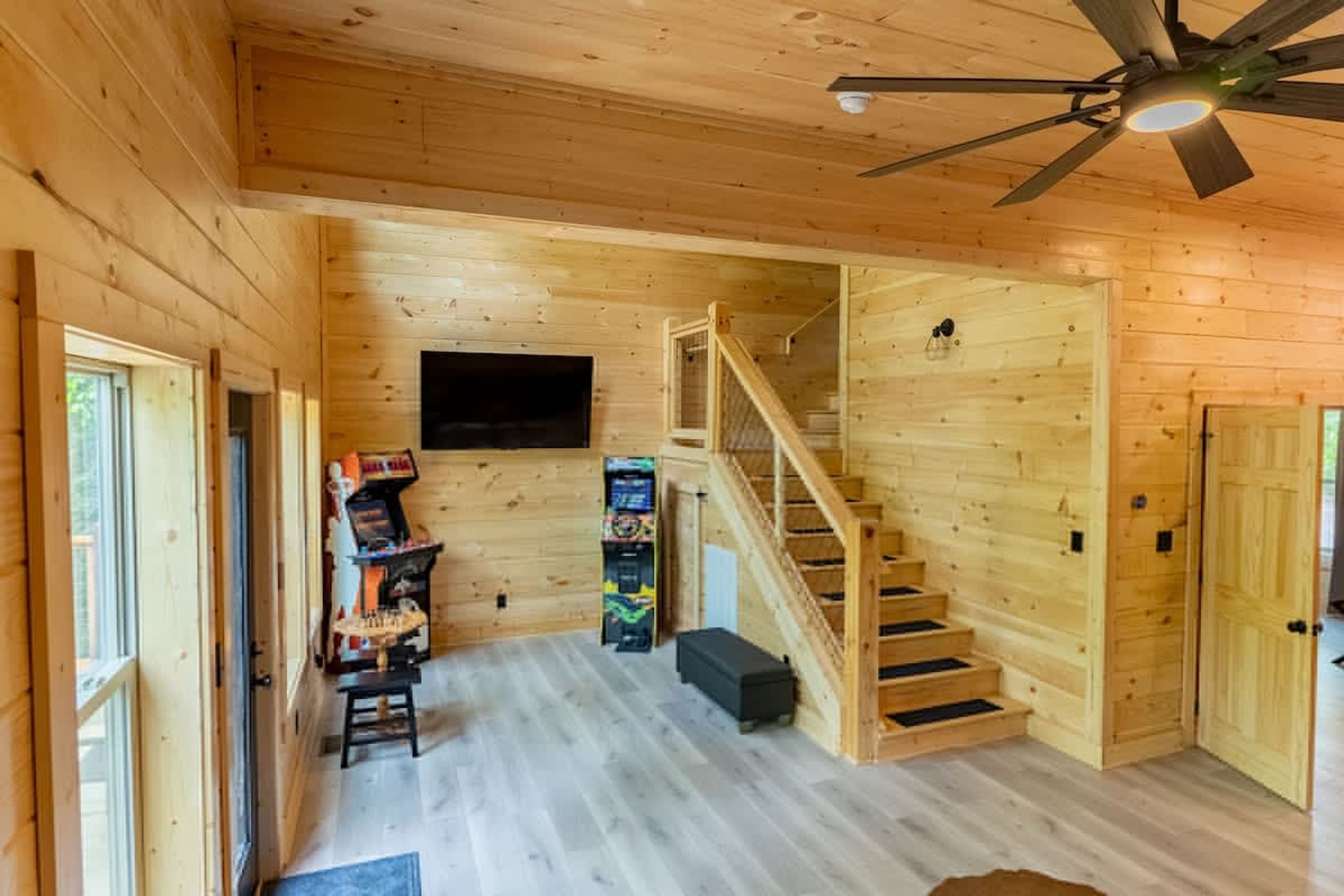 A wooden interior with a staircase, a flat-screen TV, and arcade games.