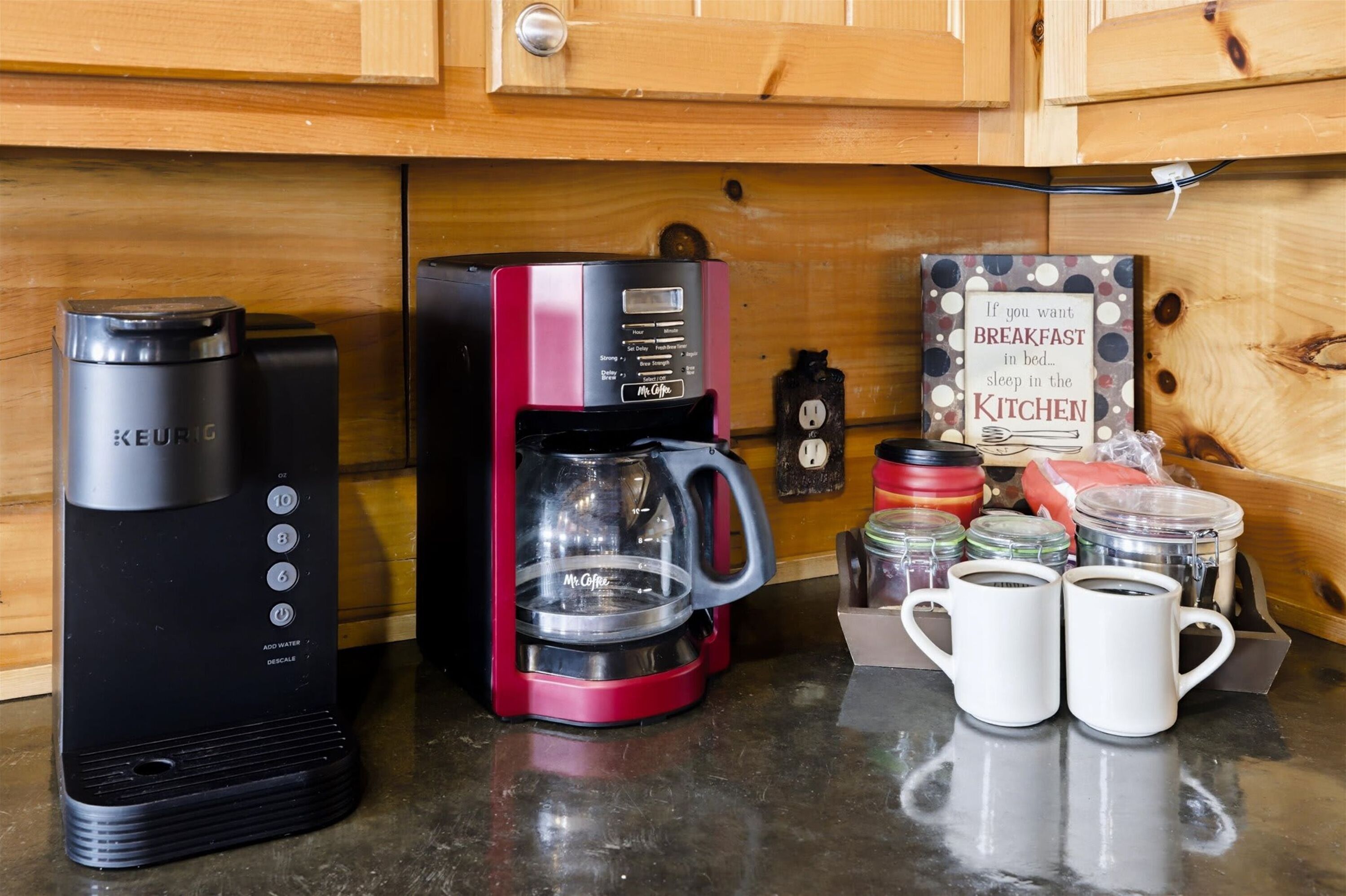 Een keuken met een Keurig koffiezetapparaat, een rode Mr. Coffee machine en een bordje met de tekst 'Als je ontbijt in bed wilt, slaap dan in de keuken'. Er staan ook twee witte mokken op het aanrecht.