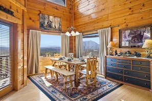 Ein Interieur einer Holzhütte mit Esstisch, Bänken, einer Chandelier, einem Schrank und einem großen Fenster mit Blick auf die Berge.