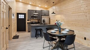 A modern kitchen with wooden walls, a central island, bar stools, and a dining table set for four. - Green Black Bear Cabins by Beyond Expectations (Sevierville)