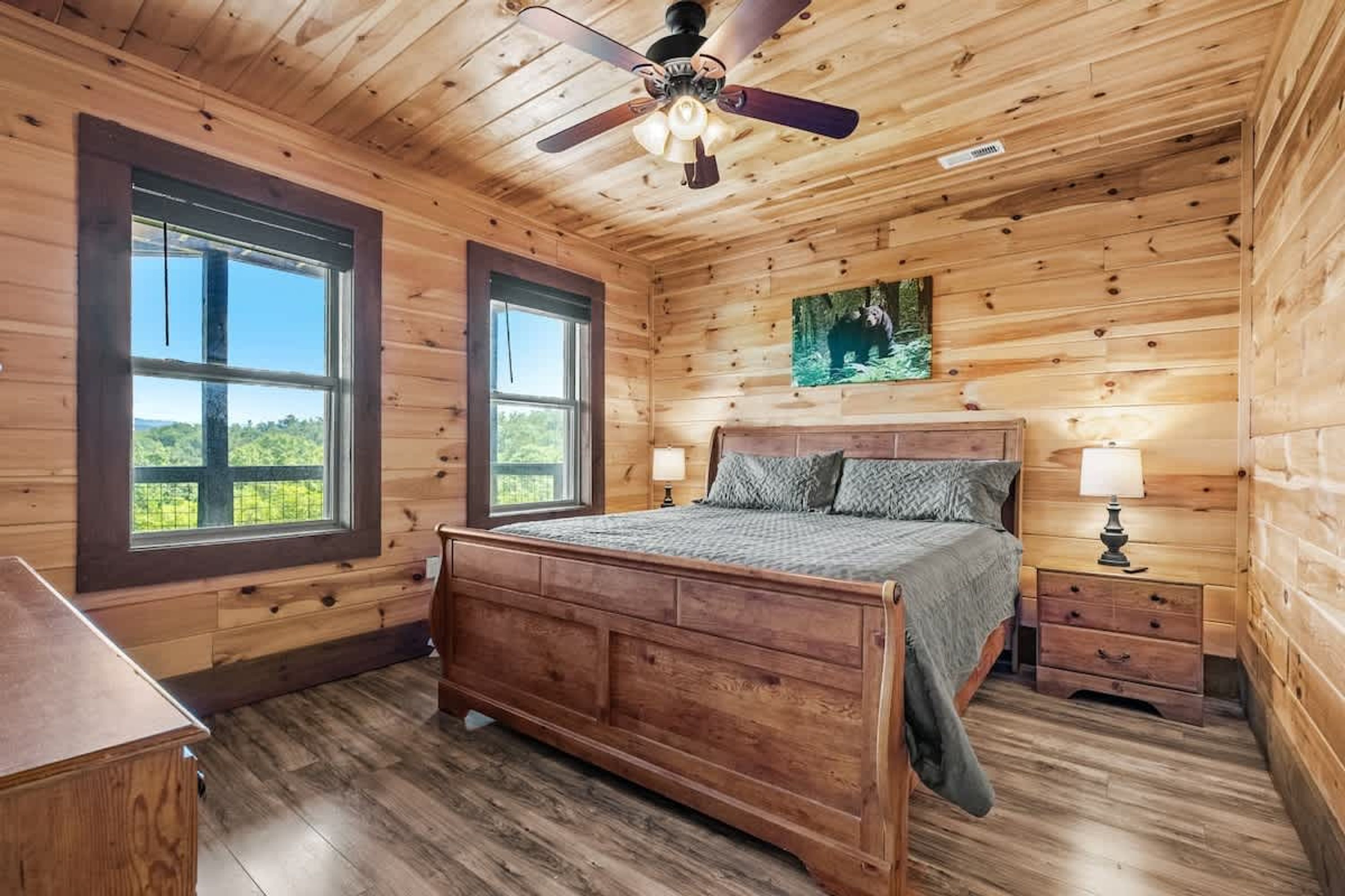 Ein Schlafzimmer einer Holzhütte mit einem Bett, zwei Fenstern, einem Deckenventilator und einem Nachttisch.