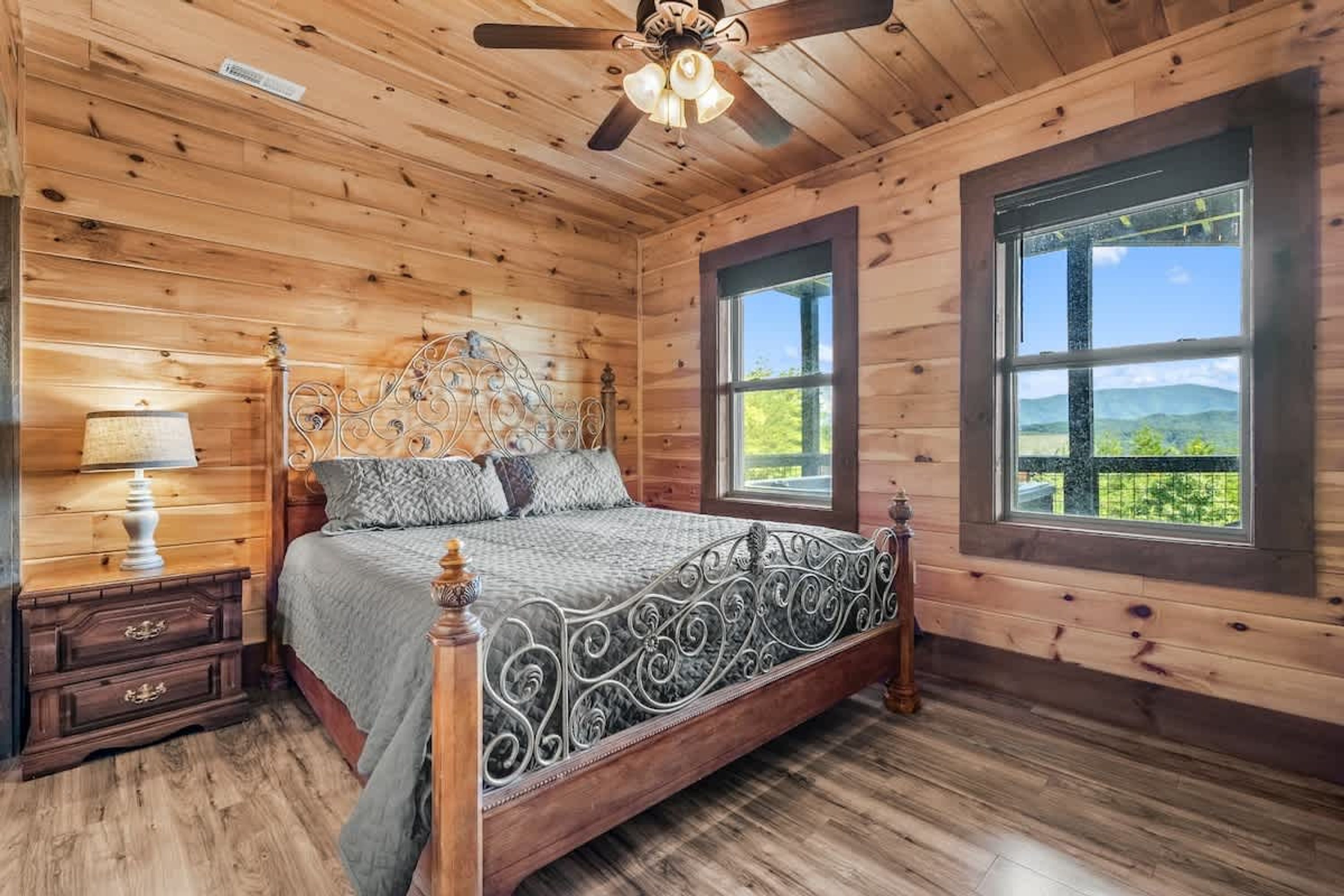 A wooden cabin bedroom with a bed, nightstand, lamp, and large windows.