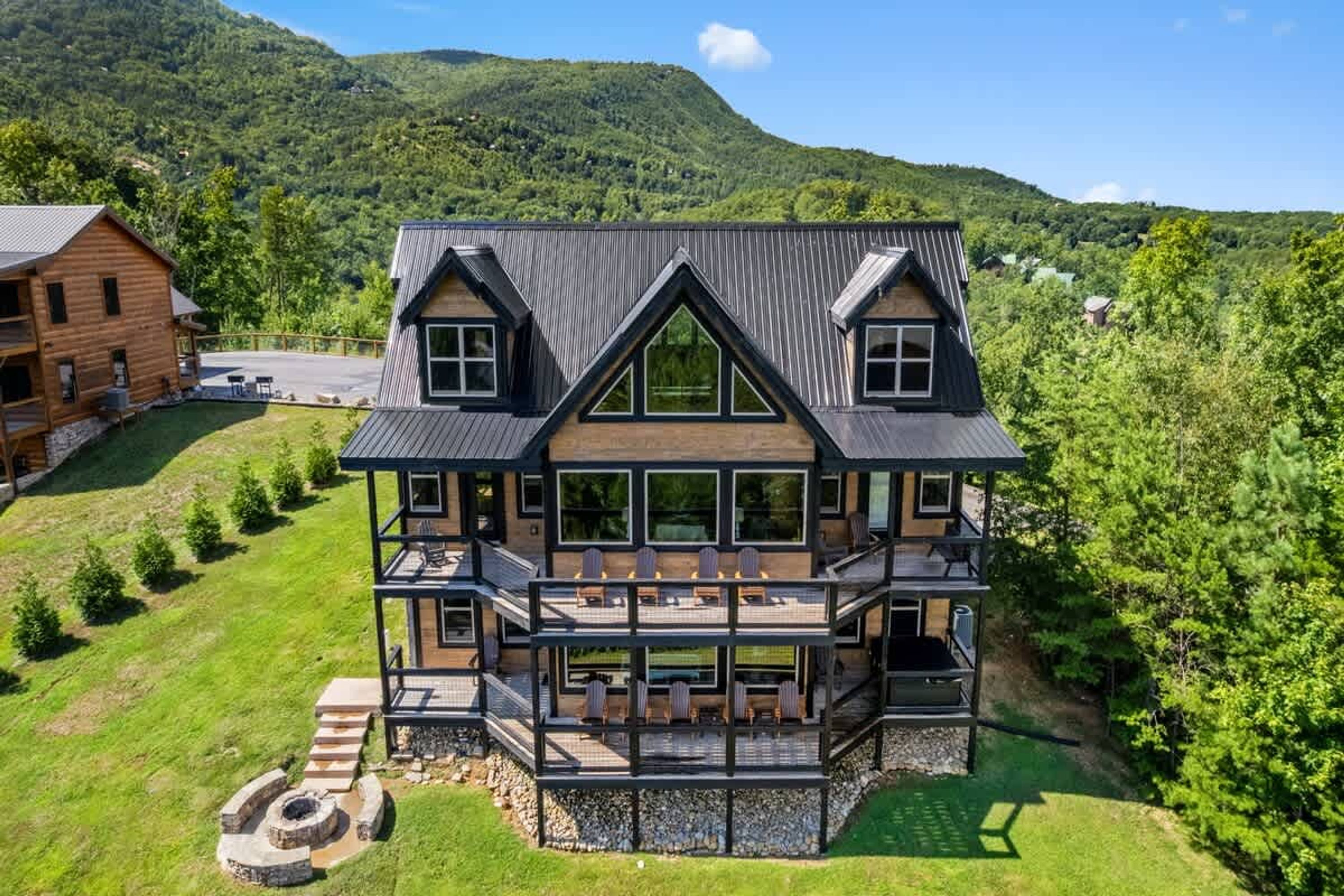 A multi-story log cabin with a balcony and outdoor seating area, surrounded by lush greenery and mountains.