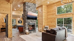 A log cabin interior with a stone fireplace, a television mounted above it, a sofa, and a large window offering a view of the outdoors. - Bearodise by Beyond Expectations With Hot Tub (Sevierville)