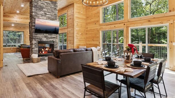 A spacious room with a stone fireplace, wooden walls, and large windows offering a view of the forest. - Bearodise by Beyond Expectations With Hot Tub (Sevierville)