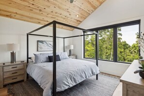 Une chambre à coucher avec un lit à baldaquin, une table de chevet avec une lampe, un bureau agrémenté d’une plante et une grande fenêtre donnant sur les arbres.