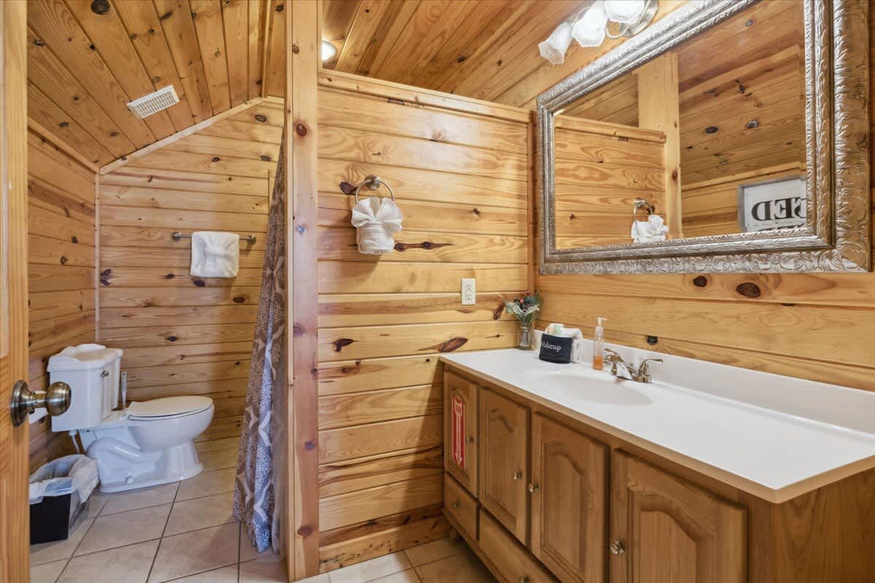 A wooden cabin bathroom with a toilet, sink, mirror, and cabinets.