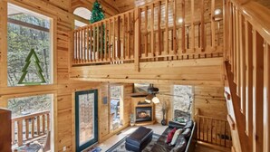 Innenansicht einer Holzhütte mit Schlafzimmer auf Schlafhöhe, Kamin, einer Couch und einem Balkon mit Weihnachtsbaum.