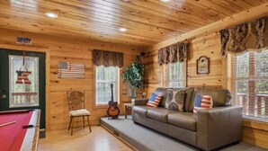 Ein Holzinnenraum mit einem Billardtisch, einer Couch mit Kissen mit amerikanischer Flagge, einer Gitarre und einer Pflanze.