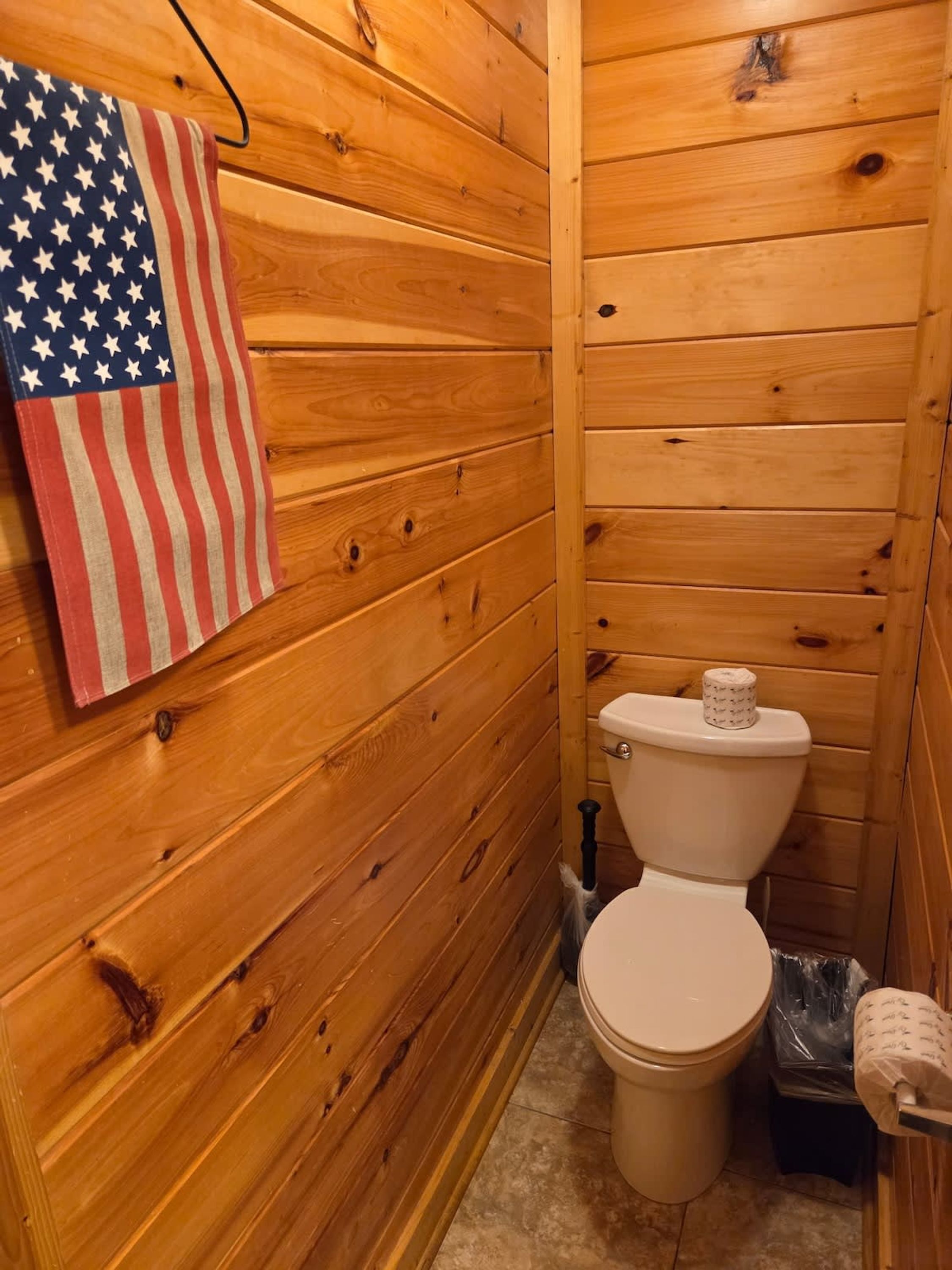 A wooden-paneled bathroom with a toilet, a trash bin, and an American flag towel.