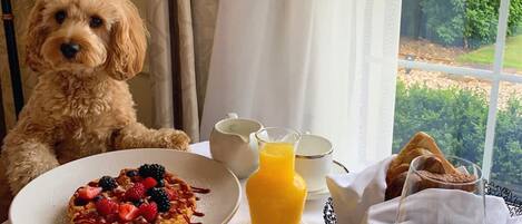 Una mesa de comedor con una porción de panqueques cubiertos de bayas, un vaso de jugo de naranja y una cesta de pan.