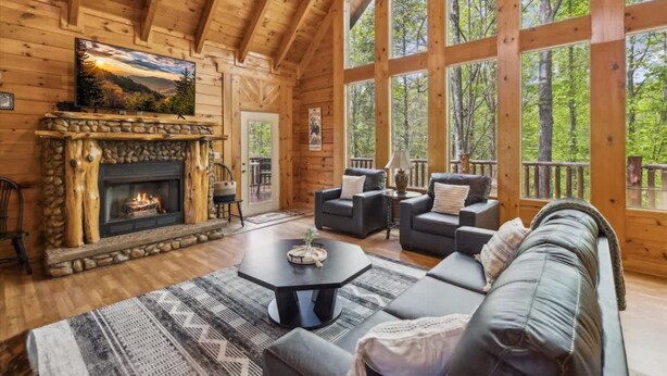 L’intérieur d’une cabane en rondins, avec une cheminée en pierre, de grandes fenêtres, un canapé et une table basse.