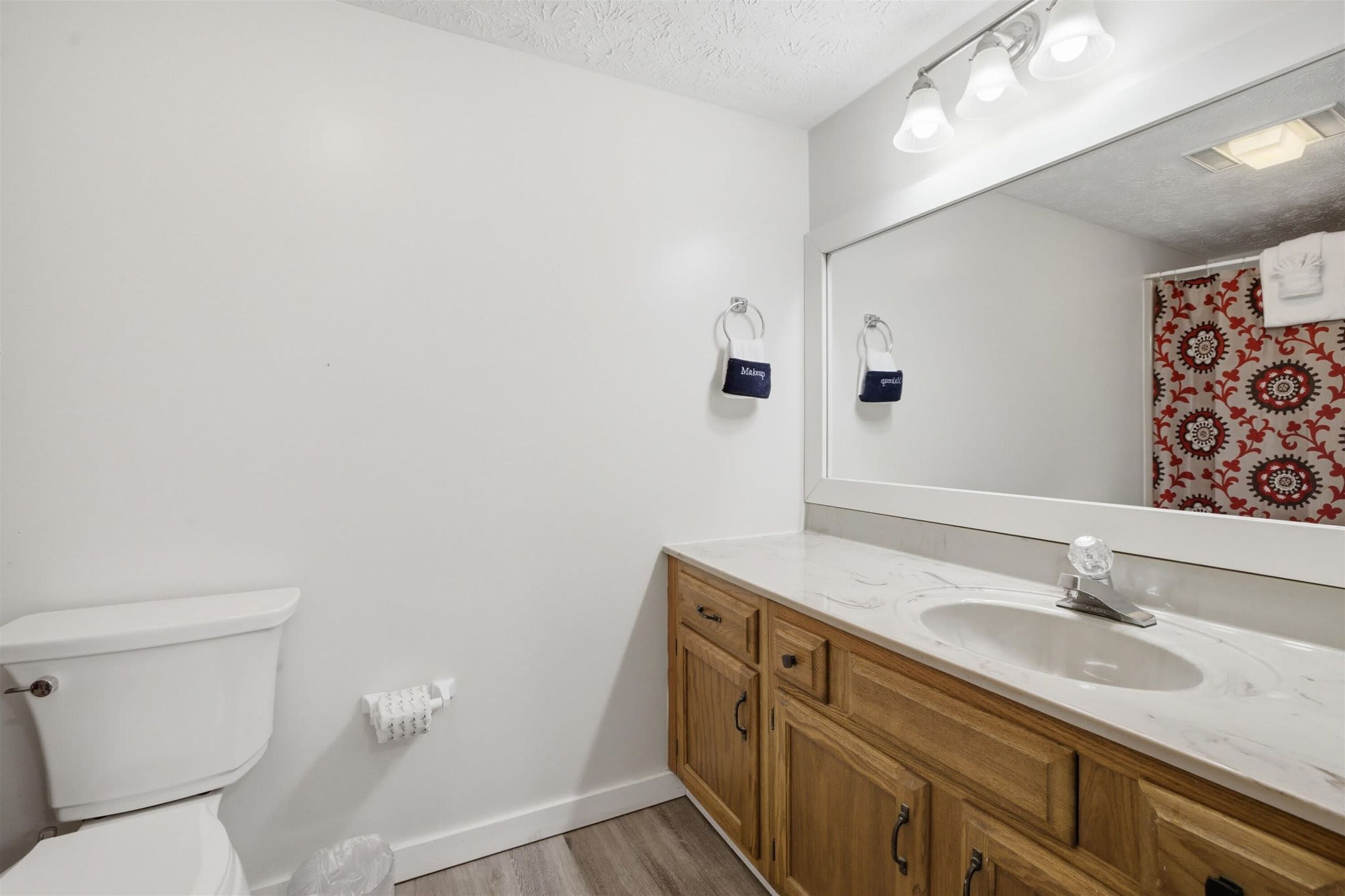 A bathroom with a toilet, a sink, wooden cabinets, a mirror, and a patterned shower wall.